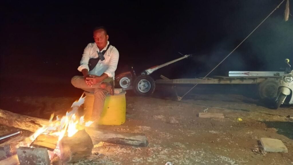 Business founder sitting in contemplation by a campfire at night, reflecting on business operations with a trailer and tractor equipment visible in the dark background.