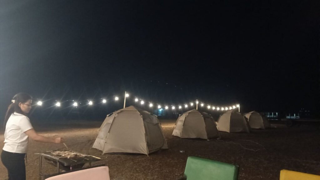 Four lighted dome tents at a camping site at night, with a string of lights above and a person grilling food on a barbecue.