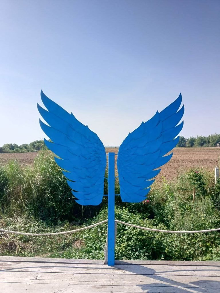 Oversized blue wooden angel wings mounted on a post at the edge of a wooden platform overlooking an open field.