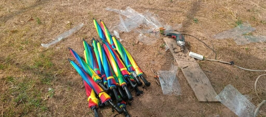 A collection of colorful rainbow umbrellas on the grass next to a bamboo pole and power tools, ready for assembly.
