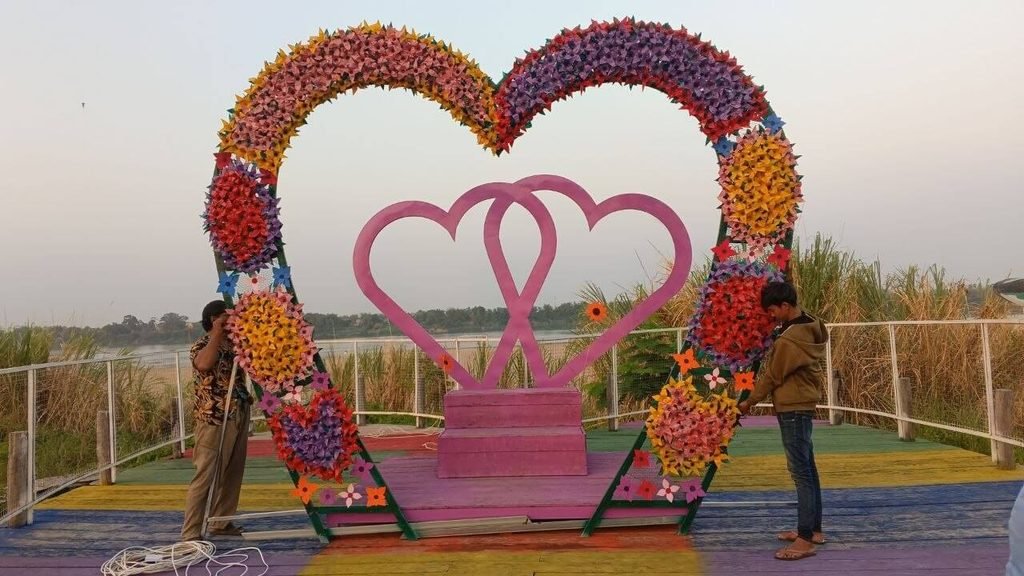 A large heart arch made of colorful paper pinwheels framing two interlocking pink hearts on a painted wooden platform.