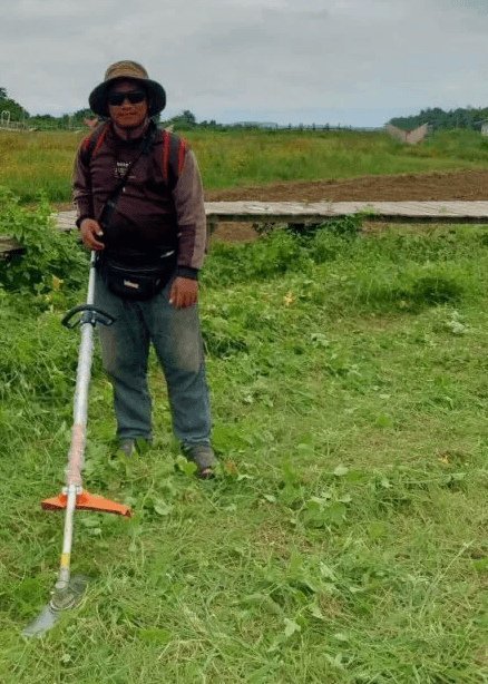 Devid Hoeur holding a grass cutter in a green field, preparing the land at Dream Garden in Kratie province.