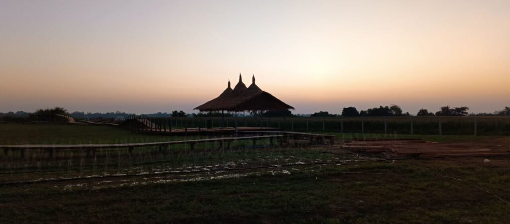 Silhouette of a handmade wooden walkway and a thatched-roof pavilion at sunset.