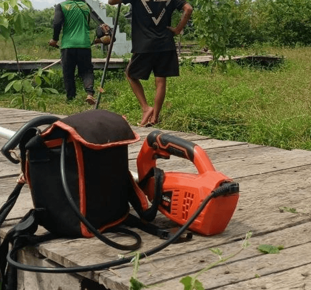 Gardening equipment resting on a wooden path with two landscaping employees using brush cutters in the background.