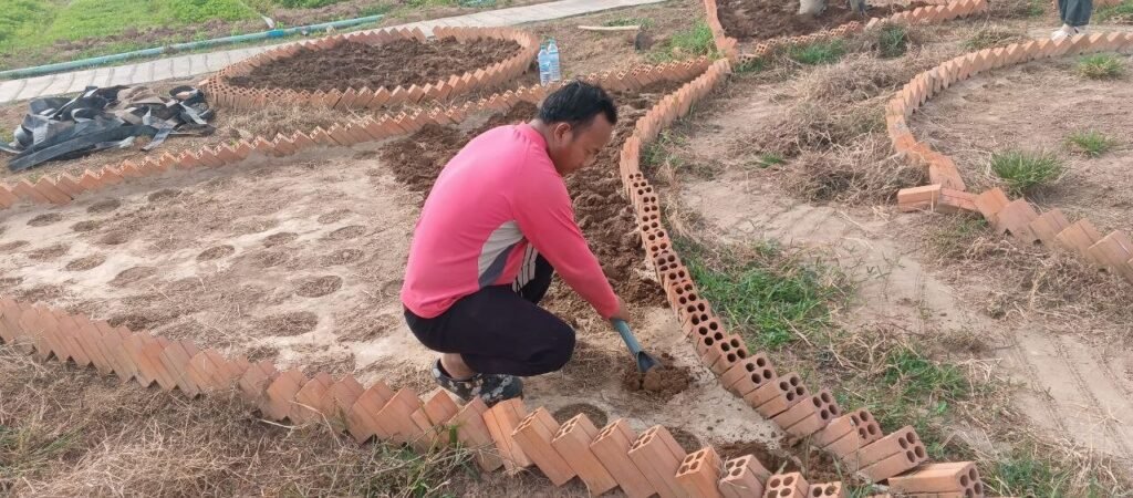 A man excavating soil and preparing planting beds inside curved brick edging under the sun.