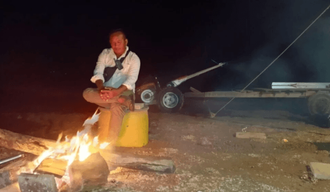 A gardener sitting alone by a campfire at night next to farm equipment, planning a second crop attempt.
