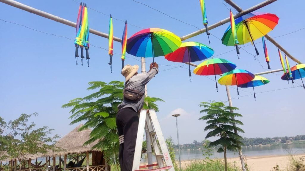 A person on a ladder secures an open rainbow umbrella to a horizontal bamboo frame using wire hooks.