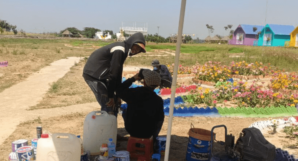 A construction site showing workers near a blue air compressor, white plastic jugs, paint buckets, and colorful garden borders in the distance.