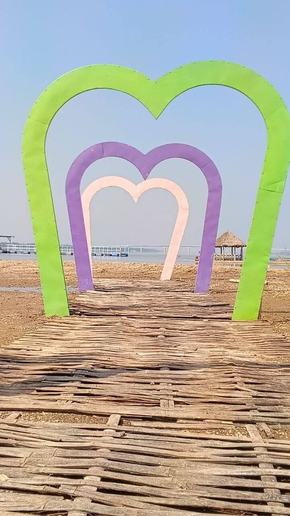 Three nested heart arches in green, purple, and pink placed along a rustic bamboo walkway leading toward an open body of water.