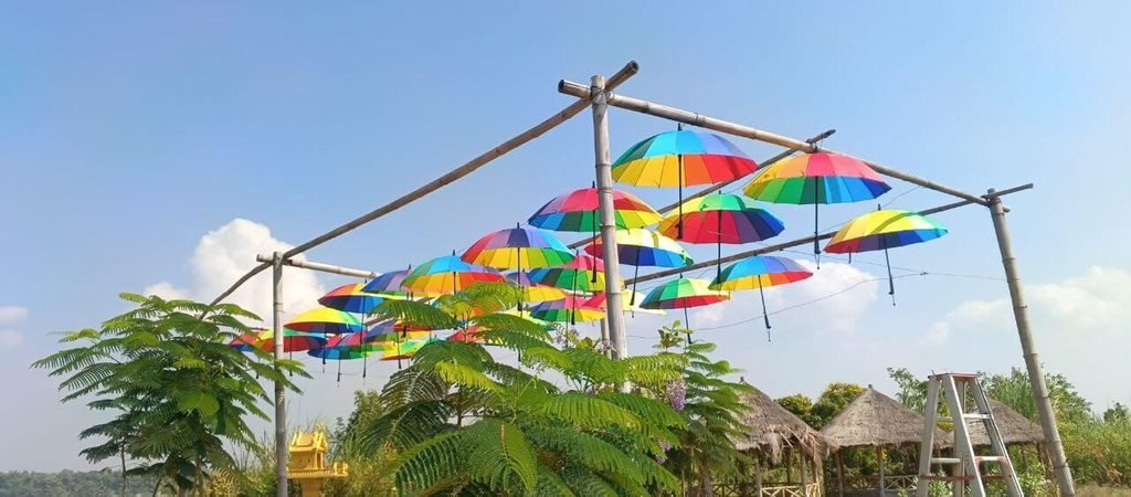 A canopy of open, rainbow-colored umbrellas suspended upside-down from a bamboo frame against a bright blue sky.