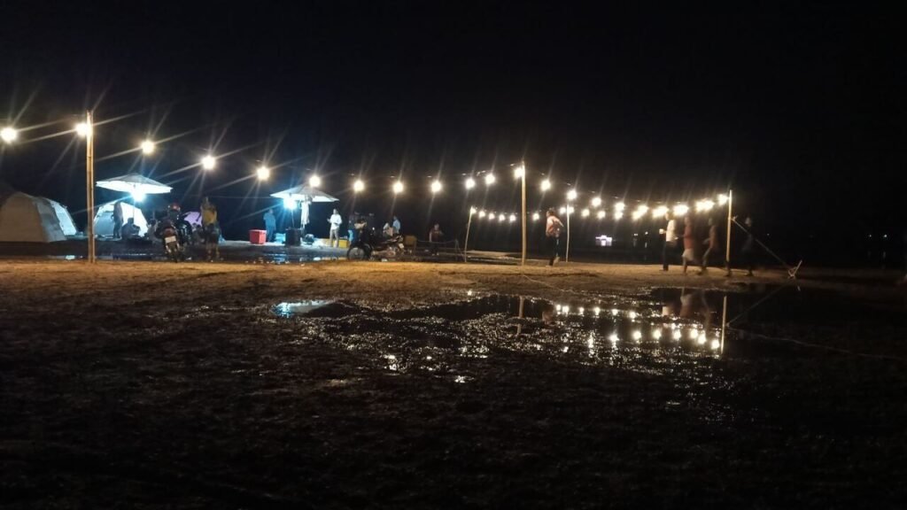 Nighttime view of a flooded outdoor camping area with string lights reflecting in large mud puddles.