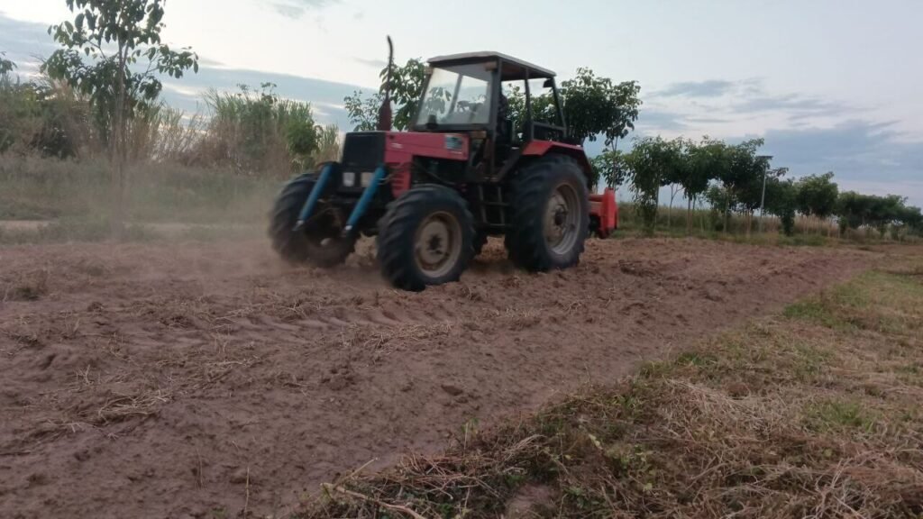 A red tractor tilling muddy, loose soil in a field during the tropical rainy season.