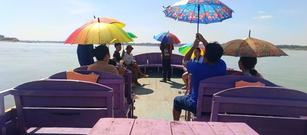 View from the back of a purple wooden boat with passengers sitting on benches and holding open, brightly colored umbrellas on a wide river under a sunny sky.