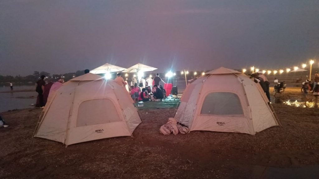 Two large camping tents set up near a river at night with lights strung up for guests.