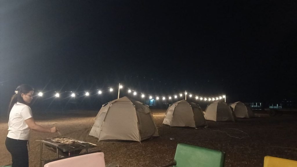 A guest barbecuing food next to canvas ground tents under string lights at a tropical glamping site.