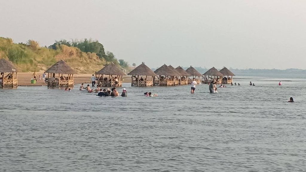 A row of bustling thatched waterside huts built directly in the shallow water with people swimming.